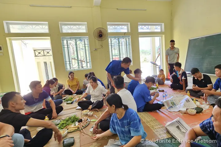 Khoảng hai mươi người ngồi trên chiếu dùng bữa trong một lớp học.