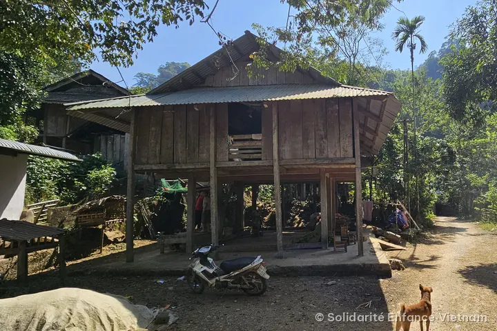 Ngôi nhà sàn bằng gỗ bên con đường đất, phía trước là một chiếc xe máy và một chú chó.