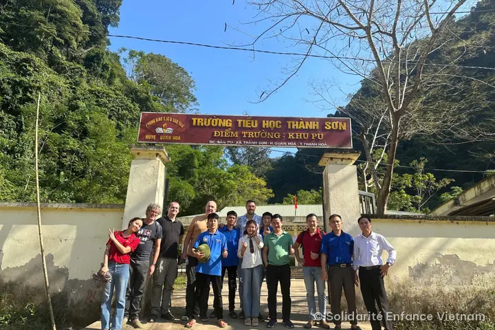 Khoảng mười lăm người chụp ảnh dưới biển “Trường tiểu học Thành Sơn – Điểm trường: Khu Pu”, dưới chân vách núi phủ rừng.