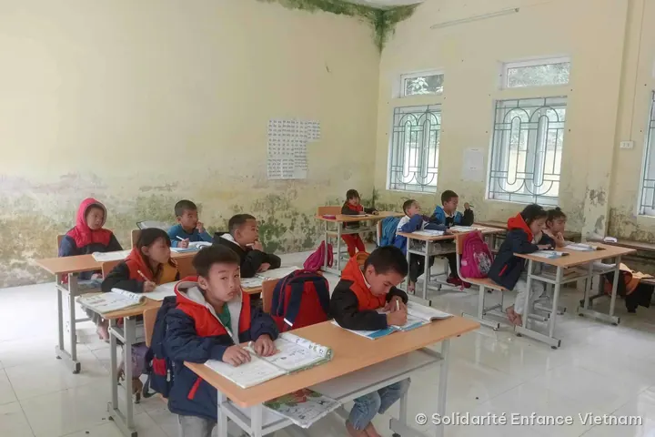 Các em học sinh mặc áo khoác đỏ đen viết bài, ngồi trên bàn gỗ sáng màu.