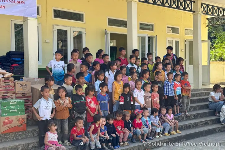 Khoảng sáu mươi em nhỏ chụp ảnh trên bậc thềm trường, bên cạnh các thùng mì, sữa, bánh và một chồng áo khoác.