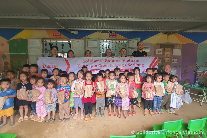 Kindergarten children holding small paper bags pose in front of the association’s banner at the Cốc Slông school.
