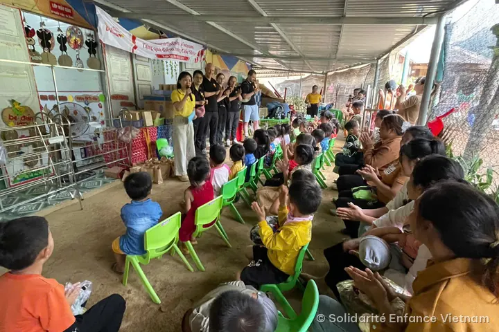 Under a shelter, children sitting on green chairs and parents applaud, facing the association’s team.