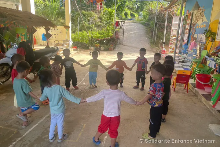 About ten young children hold hands in a circle in the yard, in front of a path leading uphill.