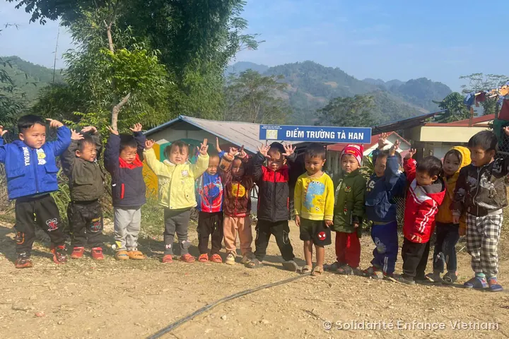 About fifteen young children in jackets pose with their arms raised in front of the “Điểm trường Tà Han” sign, with mountains in the background.