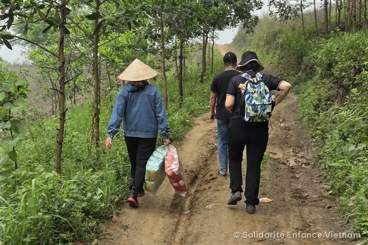 Three people seen from behind walk up a muddy dirt track between trees; one, wearing a conical hat, carries two large bags.
