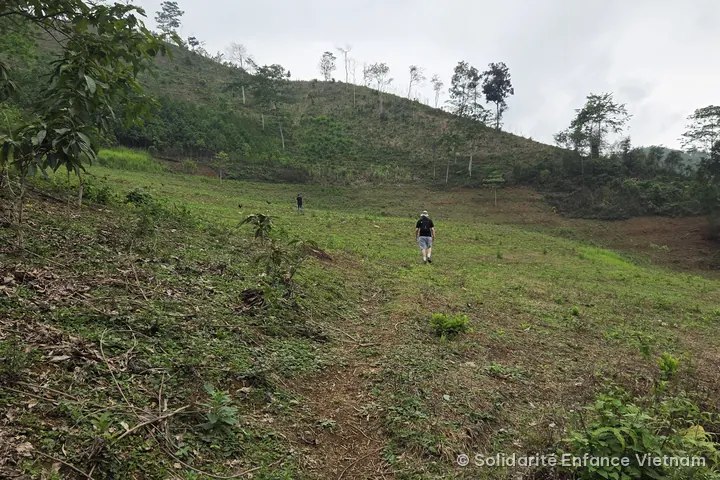 Two distant figures cross a grassy slope at the foot of a hill, under a grey sky.