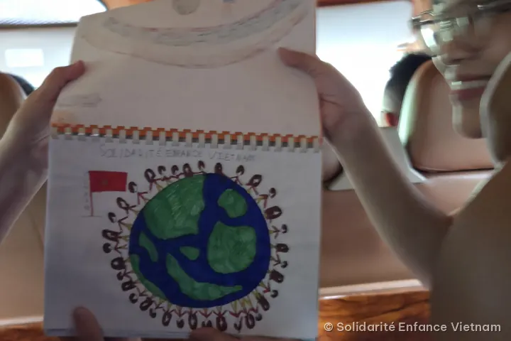Two people hold an open book showing a child’s drawing: the Earth surrounded by children holding hands, a Vietnamese flag and the words “Solidarité Enfance Vietnam”.