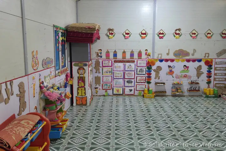 Classroom with a green tiled floor, its walls covered with cardboard decorations, children’s drawings and play corners.