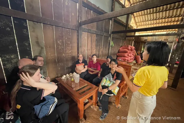 Members of the association, seated around a coffee table in a wooden house, listen to a young woman in a yellow polo shirt.
