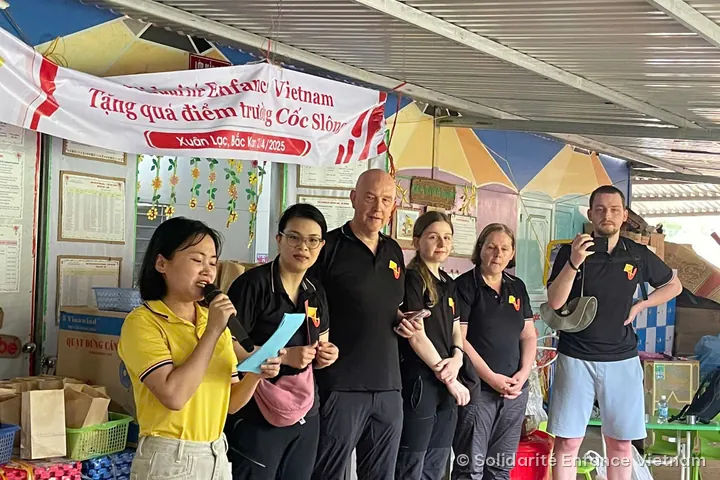 A young woman in a yellow polo shirt speaks into a microphone in front of five members of the association, under the banner “Tặng quà điểm trường Cốc Slông, Xuân Lạc, Bắc Kạn 2/4/2025”.