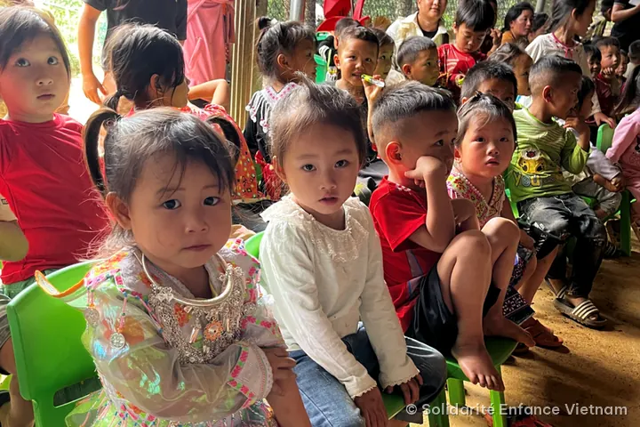 Children sitting on small green chairs look ahead, including a little girl in an embroidered traditional costume.