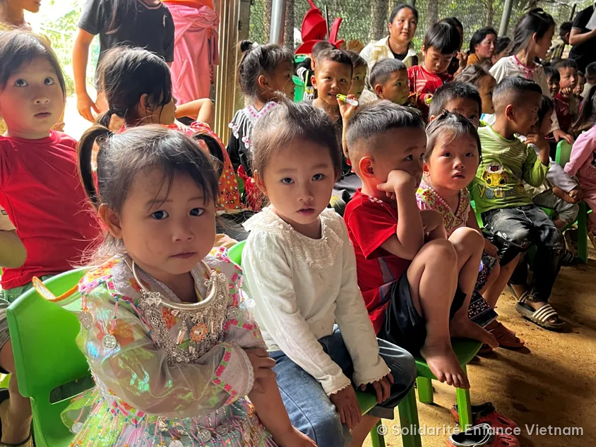 Children sitting on small green chairs look ahead, including a little girl in an embroidered traditional costume.