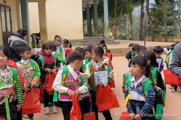Các em nhỏ, một số mặc trang phục truyền thống, đeo cặp sách xanh trên lưng và cầm túi đỏ trên tay.