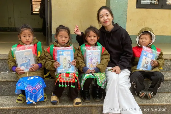 Bốn em nhỏ mặc áo khoác kaki ngồi trên bậc thềm, cầm túi đồ dùng học tập, bên cạnh một cô gái tươi cười.