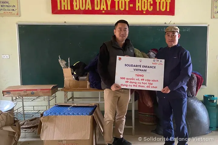 Hai người đàn ông cầm tấm bảng “Solidarité Enfance Vietnam tặng 500 quyển vở, 49 cặp sách…” trước bảng đen, xung quanh là thùng hàng và túi quần áo.