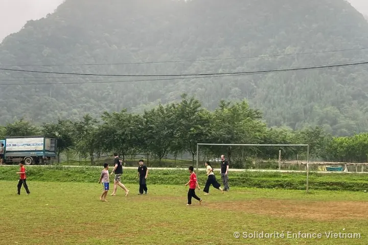 Các em nhỏ và hai tình nguyện viên đá bóng trên sân cỏ, phía sau là ngọn núi phủ rừng trong sương.