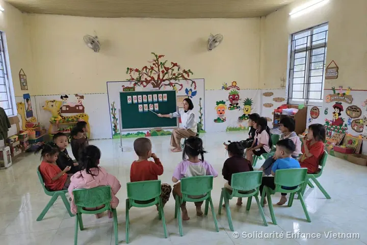Cô giáo chỉ các chữ cái trên bảng xanh cho khoảng mười hai em nhỏ ngồi thành hình vòng cung trên ghế xanh, trong căn phòng có tường được trang trí.