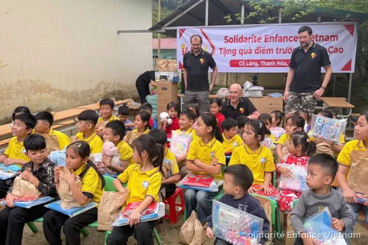 Khoảng ba mươi em nhỏ, phần lớn mặc áo polo vàng, ngồi cầm túi đồ dùng học tập, trước ba tình nguyện viên và băng rôn của hội.