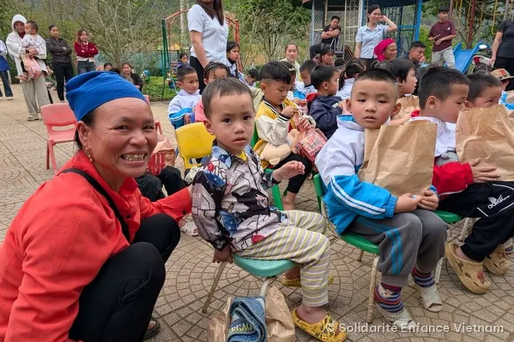 Một phụ nữ mặc áo đỏ, đội khăn xanh mỉm cười, ngồi bên các em nhỏ đang cầm túi giấy.