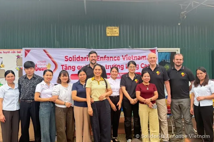 Mười bốn người lớn đứng thành hàng chụp ảnh trước băng rôn của hội và một dãy nhà màu xanh.