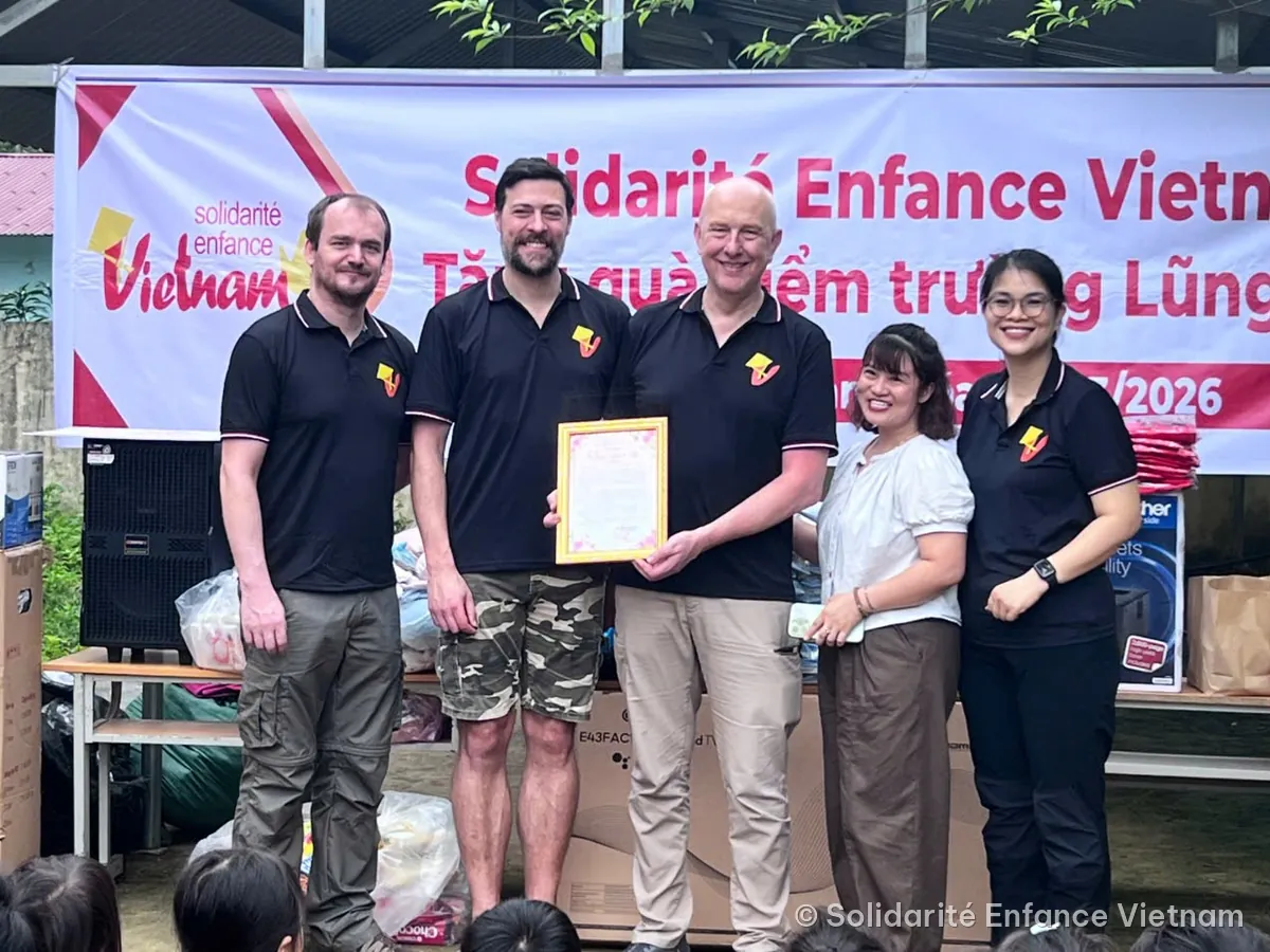 Five people smile in front of the association’s banner, holding a framed document, surrounded by boxes of supplies.
