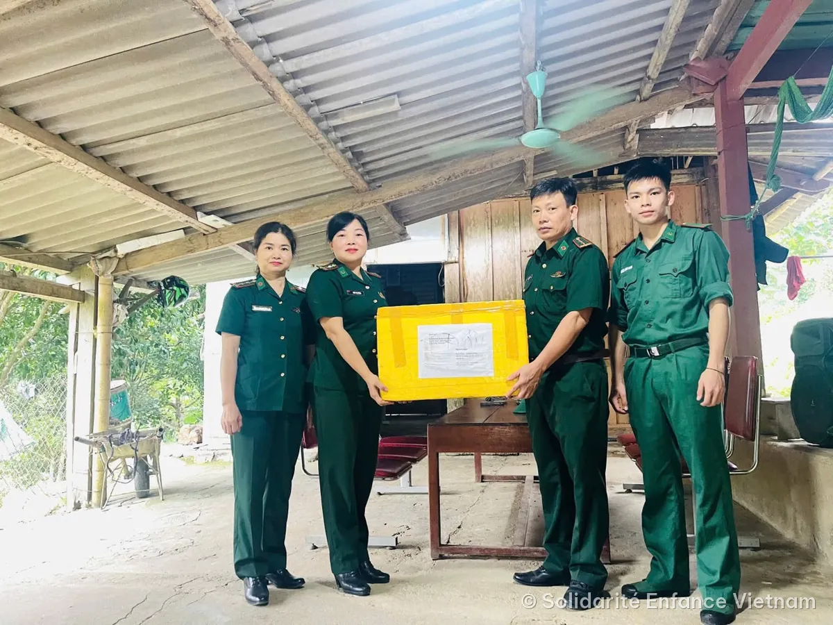 A box of medicines is handed over to people in green uniforms, under a canopy.