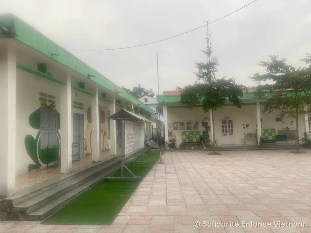 Sân lát gạch của trường mầm non Tân Quang, xung quanh là các dãy nhà trắng mái xanh; một chú ếch được vẽ trên tường.