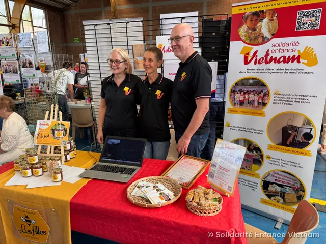 Trois bénévoles en polo noir de l’association posent devant un stand de vente de miel, à côté de l’affiche de présentation.