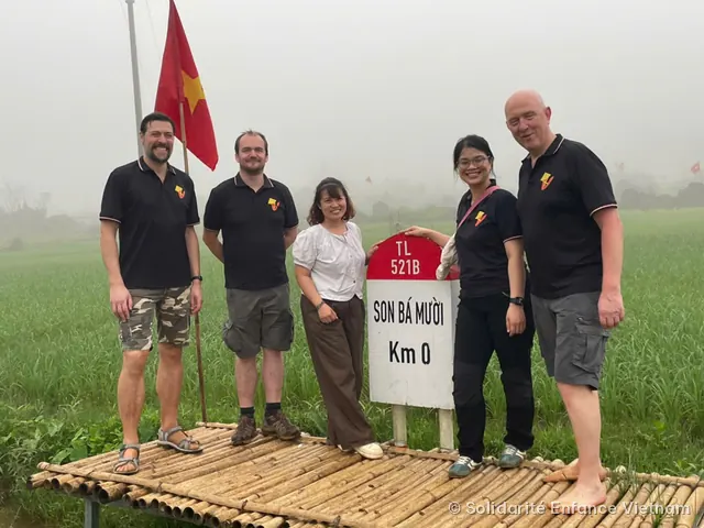 Cinq personnes, dont quatre en polo noir de l’association, posent sur une plateforme en bambou devant une borne kilométrique et un drapeau vietnamien.